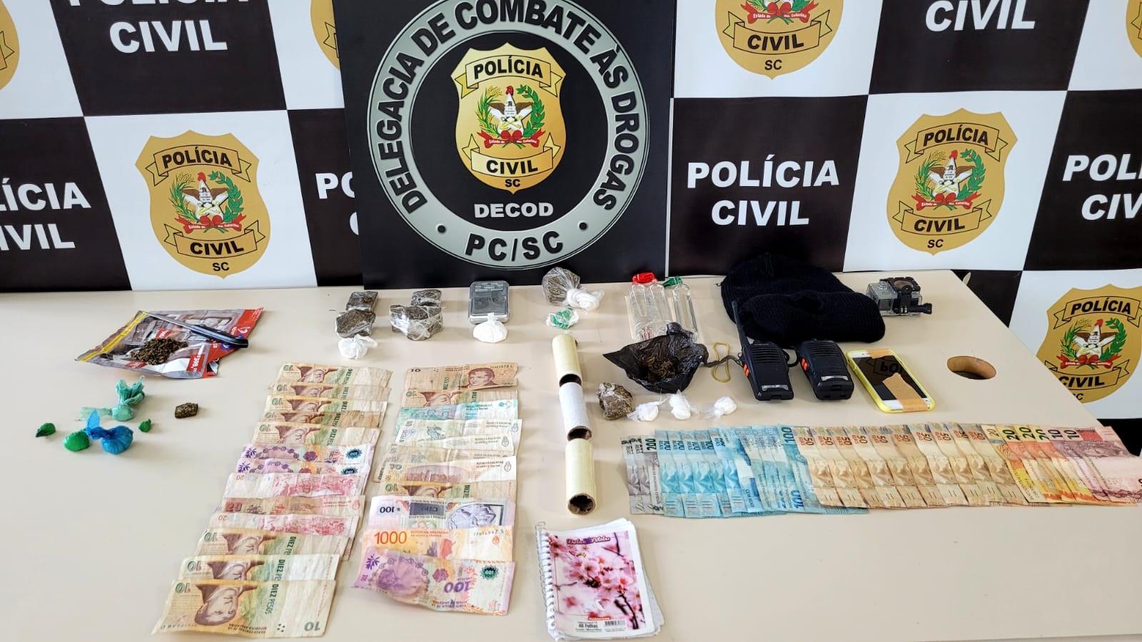 sobre a mesa dinheiro, drogas (maconha, cocaína e lança perfume), dinheiro proveniente do tráfico (notas picadas), balança de precisão, rádios comunicadores e agenda com anotações da contabilidade do tráfico