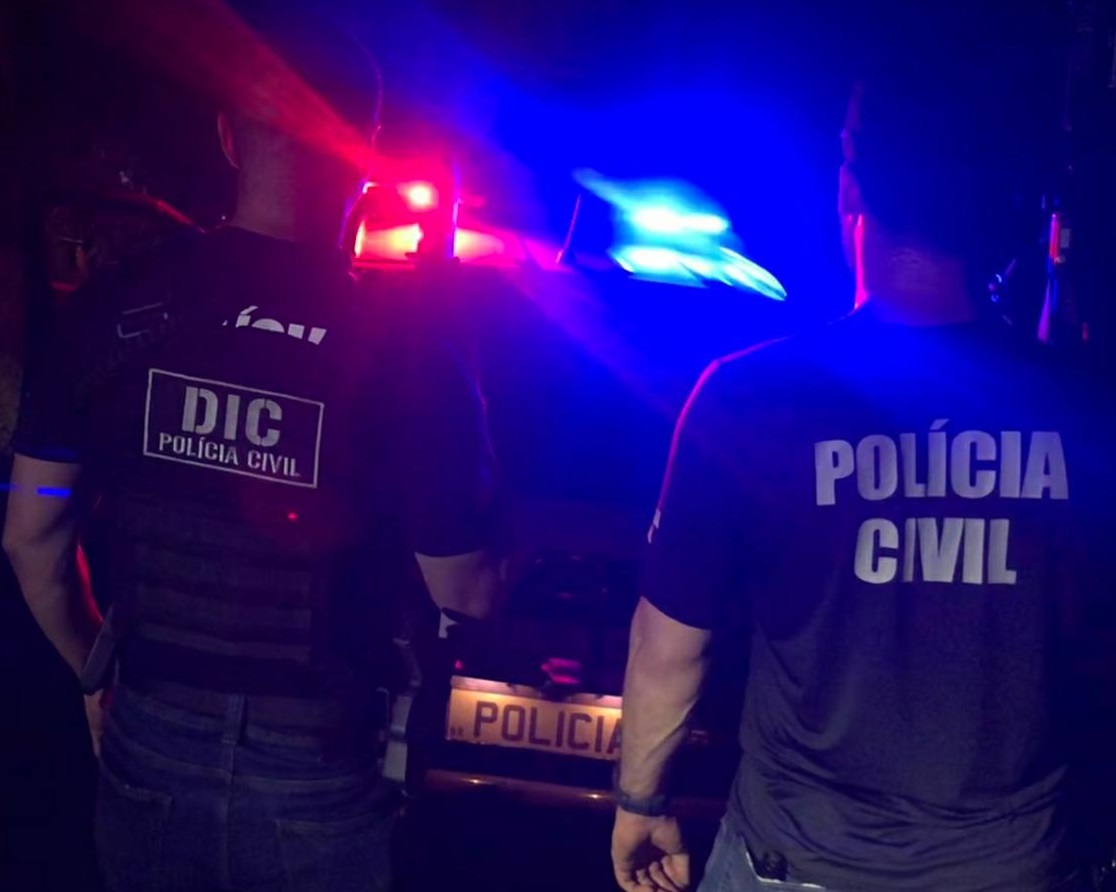 POLICIAIS EM FRENTE A UMA VIATURA COM A CIRENE LIGADA, O DA ESQUERDA ESTÁ COM A ROUPA COM O DIZER DIC POLICIA CIVIL E O DA DIREITA POLICIA CIVIL