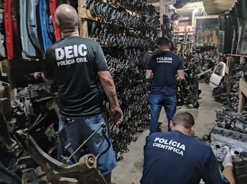 dois policiais da policia cientifica e um policial civil próximo a parte de carros desmontados nas paredes