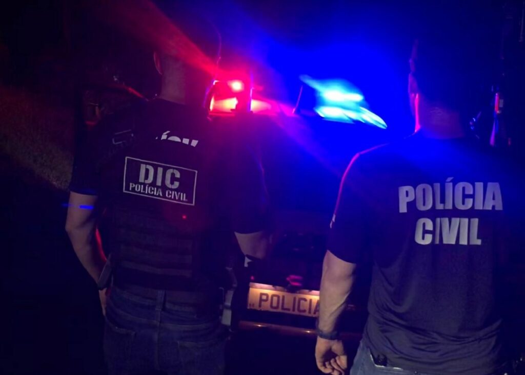 POLICIAIS EM FRENTE A UMA VIATURA COM A CIRENE LIGADA, O DA ESQUERDA ESTÁ COM A ROUPA COM O DIZER DIC POLICIA CIVIL E O DA DIREITA POLICIA CIVIL