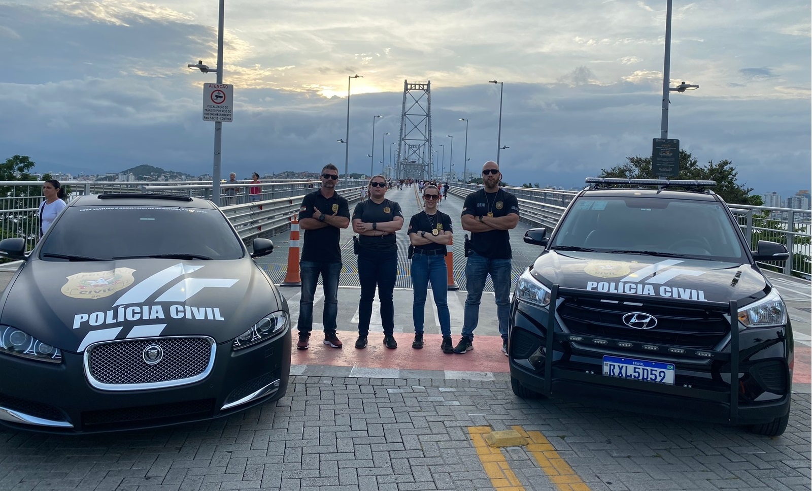 Ponte Hercílio Luz de fundo, na frente quatro policiais e duas viaturas da Polícia Civil