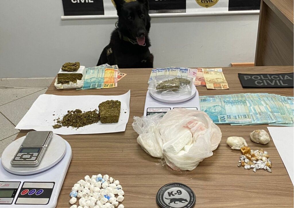 CÃO DA OPERAÇÃO, NA FRENTE MESA COM DROGAS, DINHEIRO, BALANÇA E LOGO DA OPERAÇÃO DE POLICAIS COM CÃES