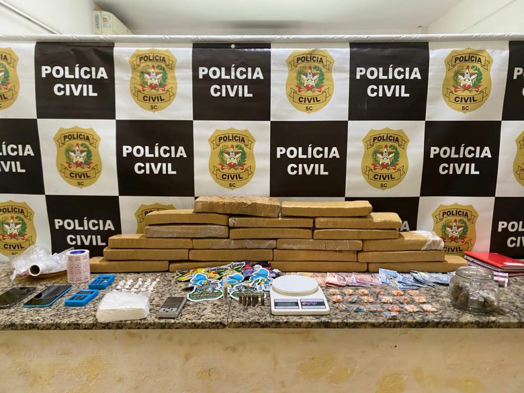 MESA EM FRENTE A UM BANNER, NA MESA DROGAS, CALCULADORAS, TELEFONES, BALANÇA, DINHEIRO, ADESIVOS E SACOLAS