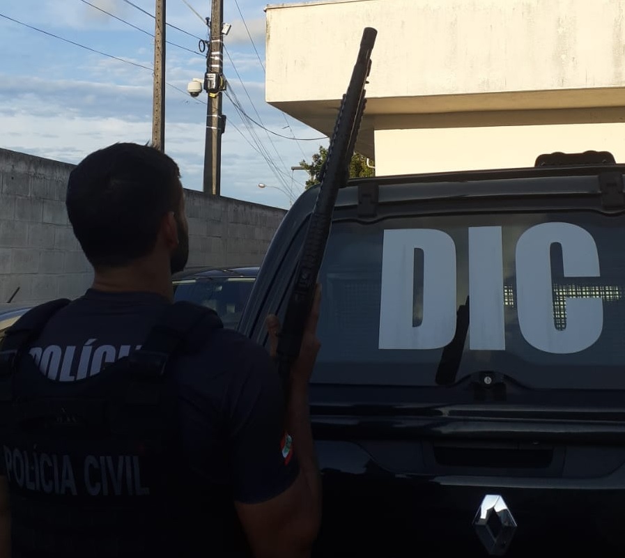 policial segurando uma arma em frente a uma viatura da DIC