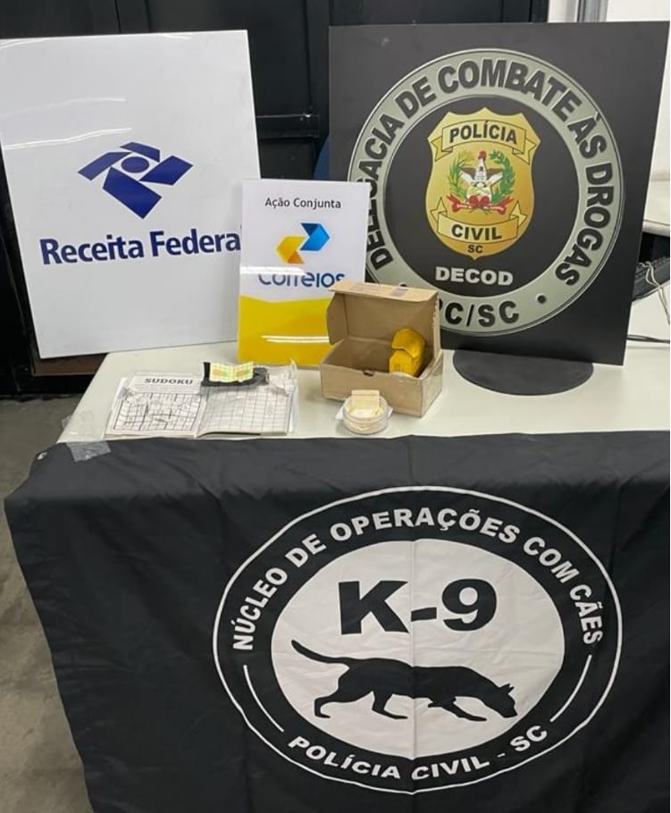mesa com banner da Receita Federal, Correios e Delegacia de Combate às drogas junto a estes um pacote e faixa Coordenadoria de Operações Policiais com Cães