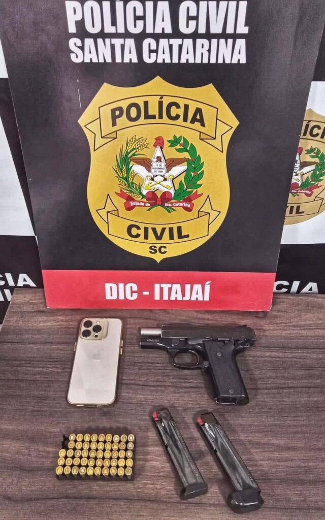 POSTER DA DIC DE ITAJAI JUNTO NA MESA CELULAR, ARMA E MUNIÇÕES