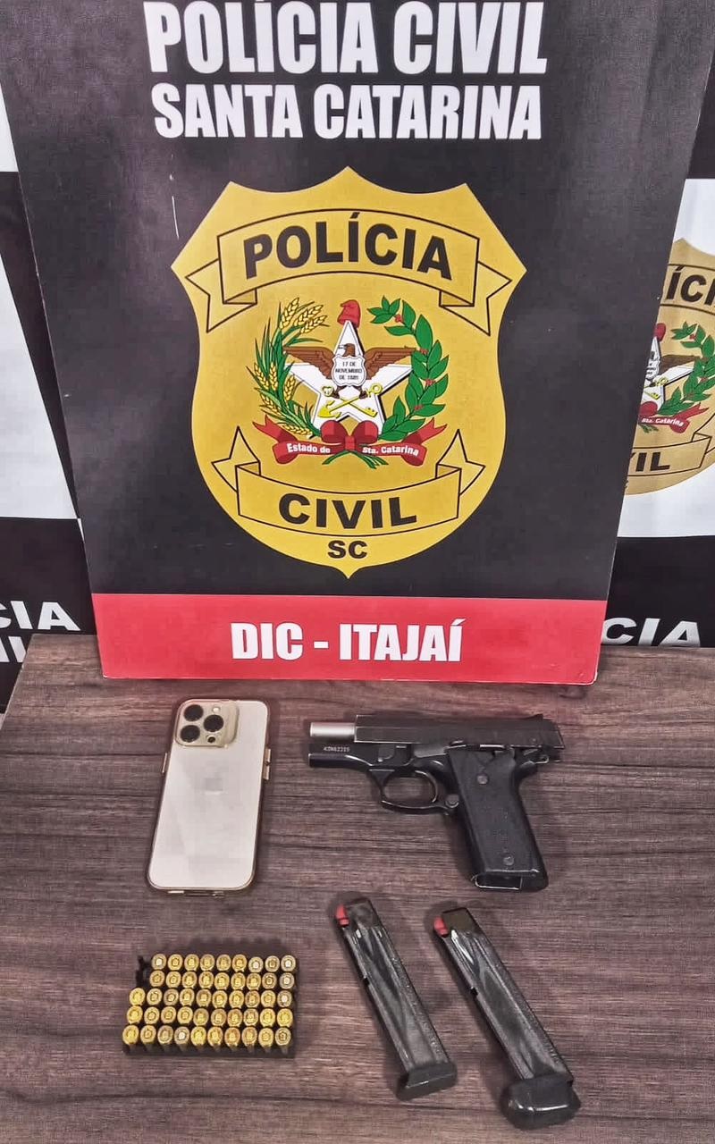 POSTER DA DIC DE ITAJAI JUNTO NA MESA CELULAR, ARMA E MUNIÇÕES