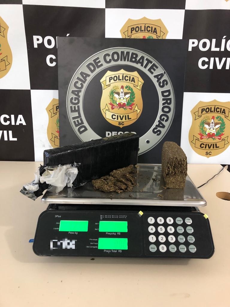 banner atrás, a frente e sobre uma balança cartaz da Delegacia de Combate as Drogas e dois tabletes de droga