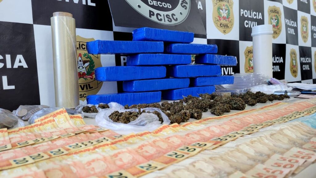 banner ao fundo na mesa poster da delegacia da Polícia Civil de combate as drogas com drogas, dinheiro, cartões, celulares, facas e embalagens recolhidas