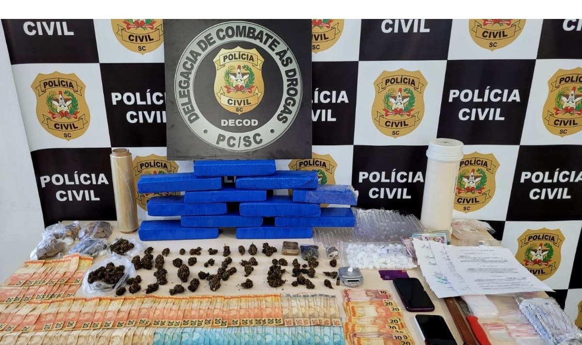 banner ao fundo na mesa poster da delegacia da Polícia Civil de combate as drogas com drogas, dinheiro, cartões, celulares, facas e embalagens recolhidas