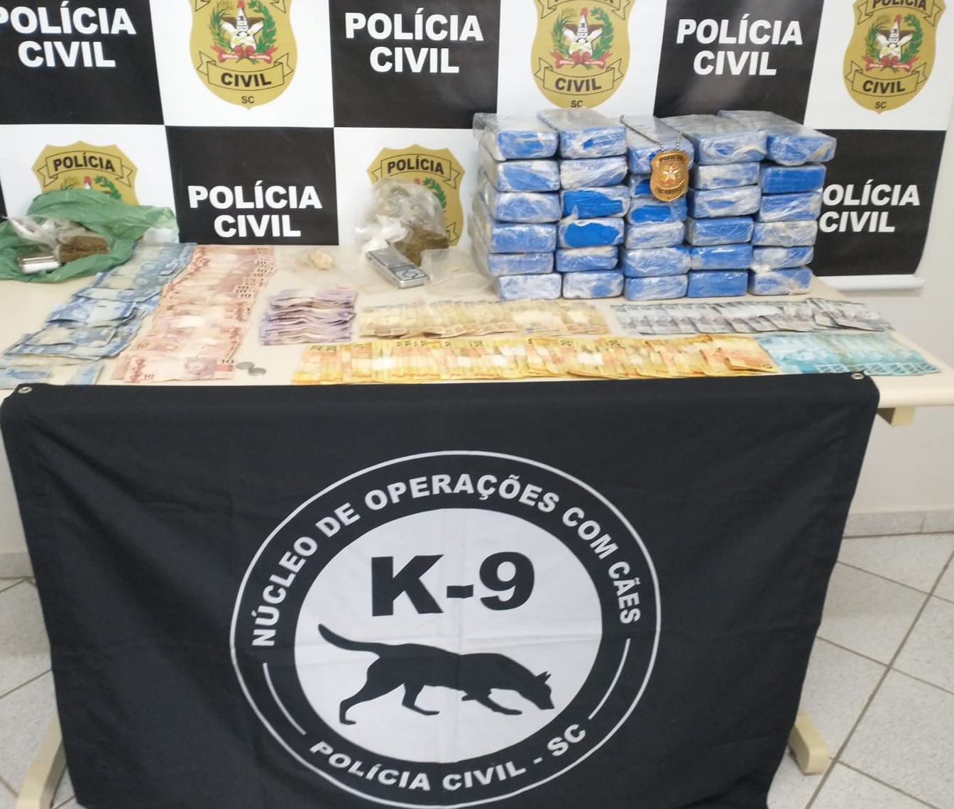 banner da policia civil de fundo encima da mesa dinheiro, tabletes de droga e a frente deste uma insígnia da policia civil , balança, sacolas e a baixo um manto do poster do Núcleo de Operações com Cães da Policia Civil