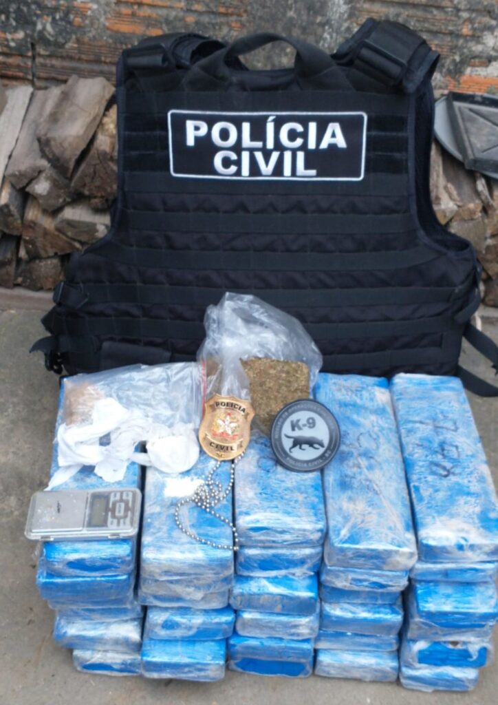 muro de tijolos e a frente deste colete da Policia Civil junto com tabletes de droga e a insígnia da Policia Civil e do Núcleo de Operações com Cães da Policia Civil