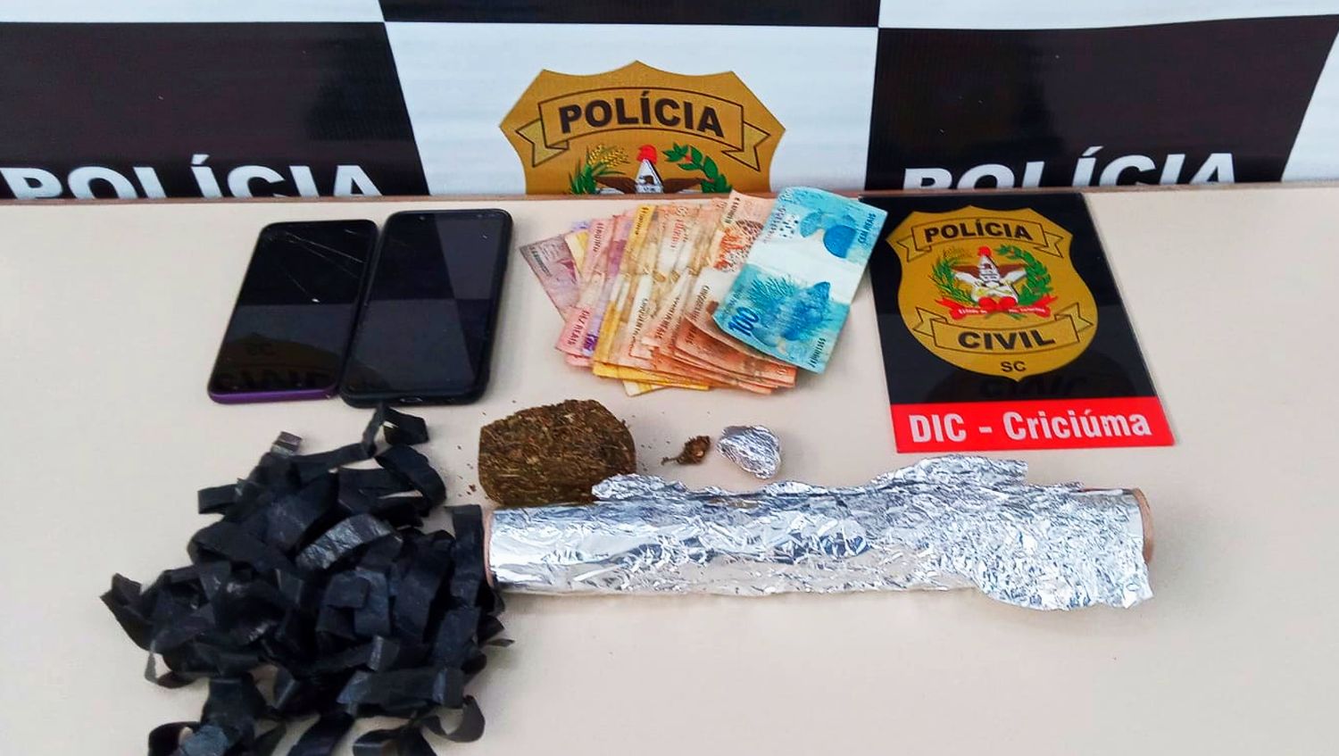 dinheiro, dois celulares, fita de carbono e rolo de papel alumínio sobre uma mesa ao lado de um pôster da DIC de Criciúma