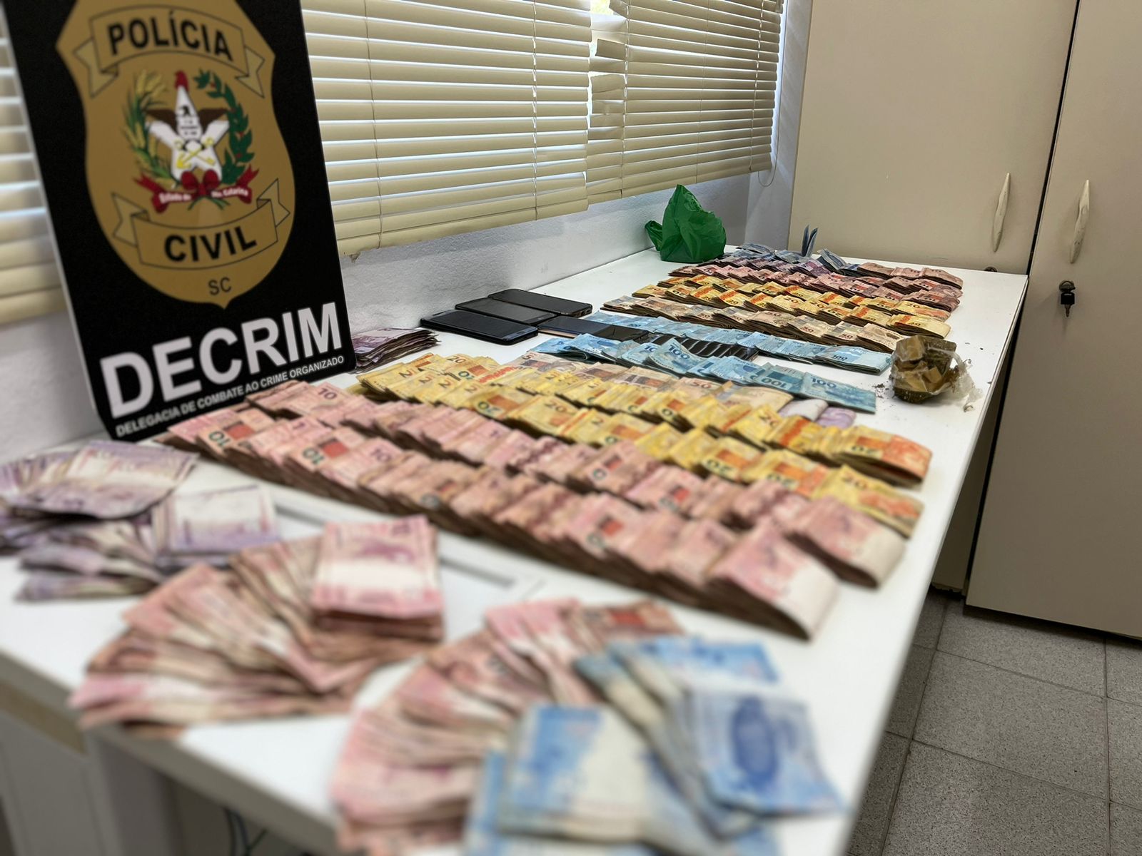 Poster da DECRIM, dinheiro, 5 celulares e droga sobre uma mesa